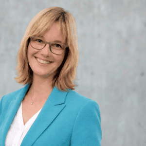 Professionelle Frau in blauer Jacke und Brille lächelnd vor neutralem Hintergrund.