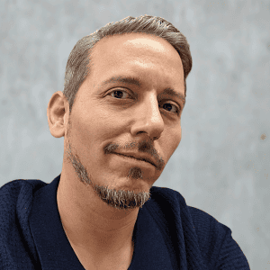 Porträt eines Mannes mit kurzen, grauen Haaren und Bart, lächelnd vor neutralem Hintergrund.